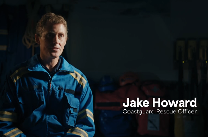 A coastguard rescue officer in blue overalls sits against a dark backdrop