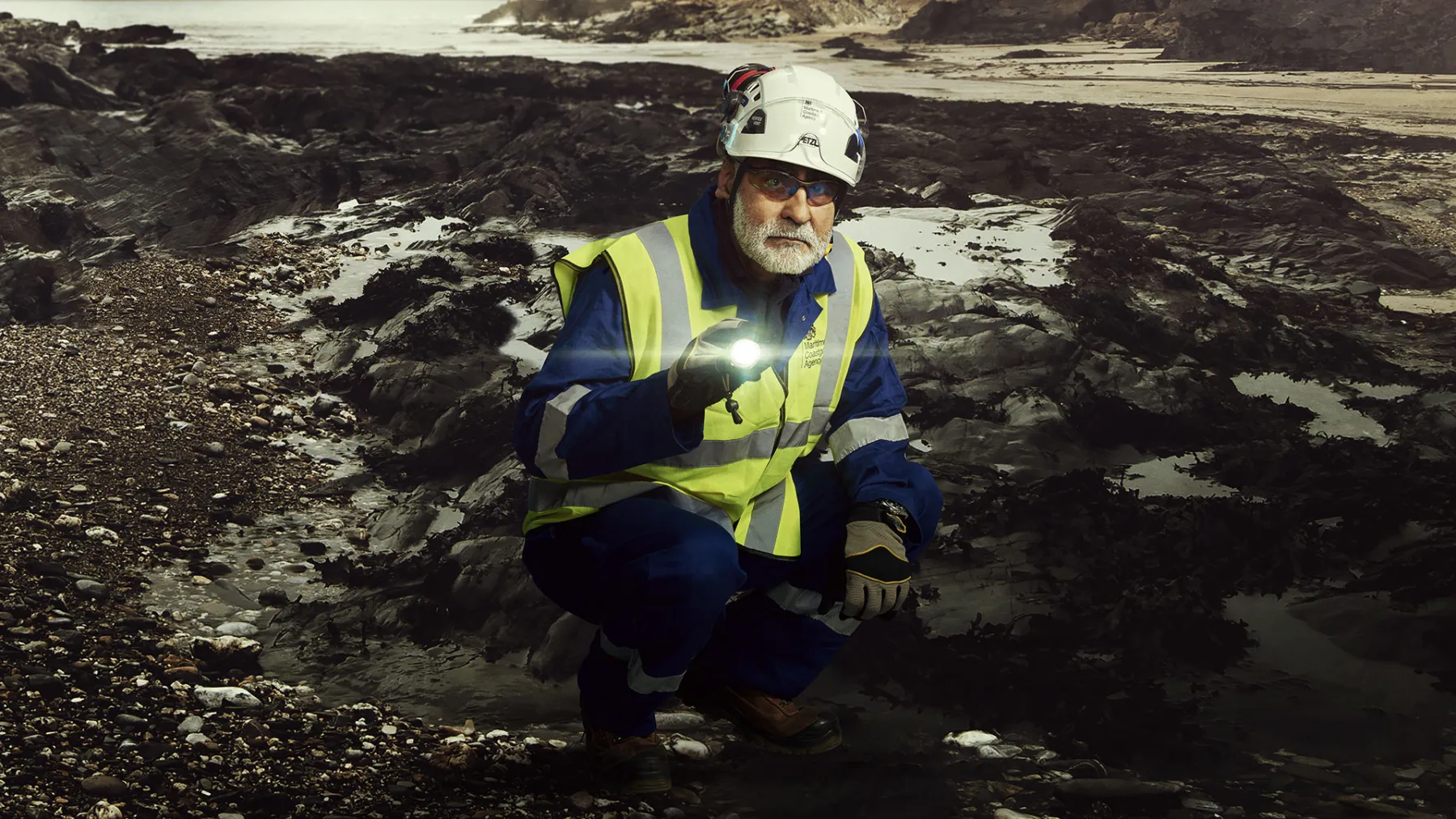 A man wearing a hard hat and high-vis jacket crouches on a rocky landscape. he is holding a torch and looking intensely at the camera.