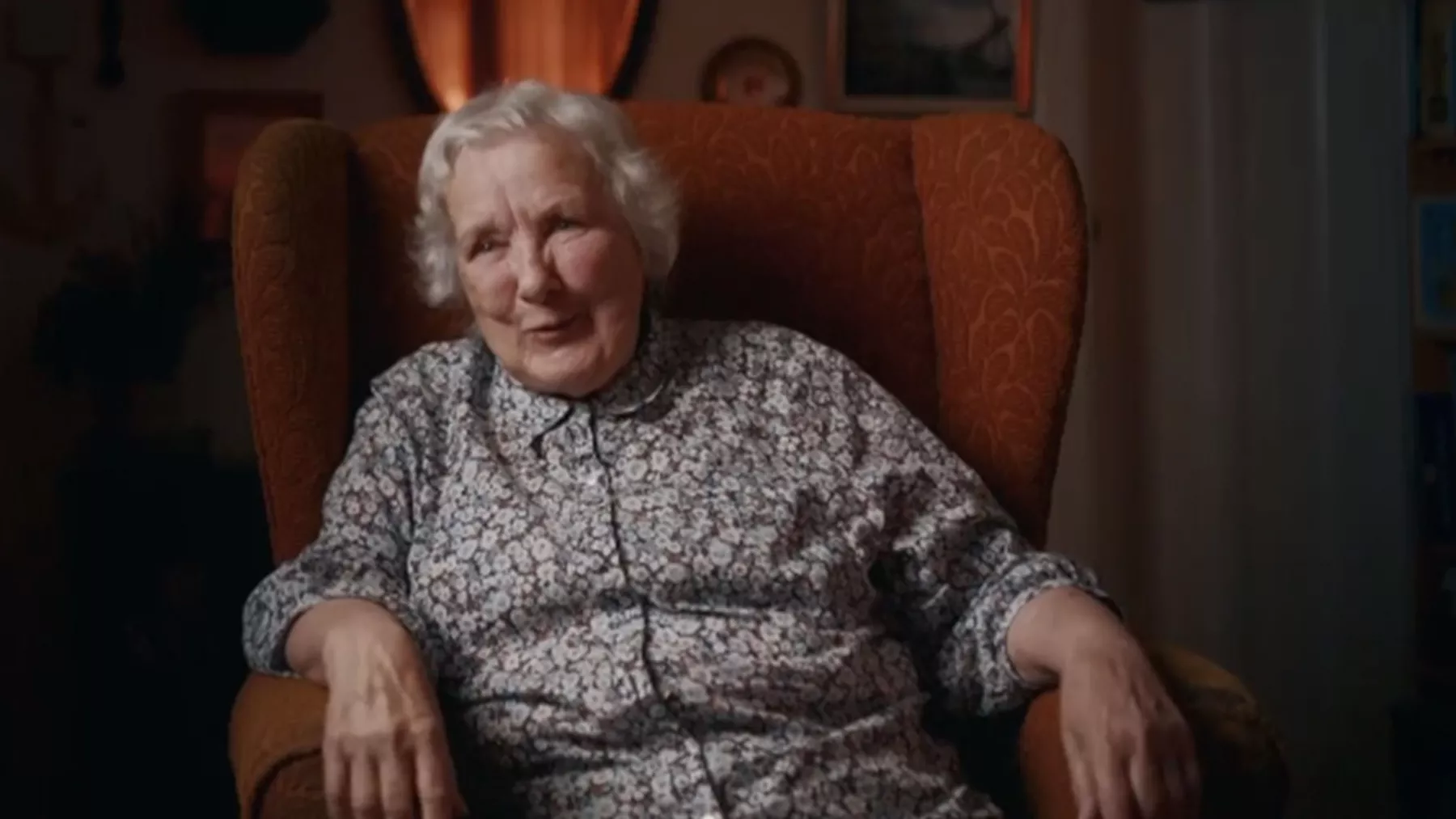 A lady with grey hair sits in an armchair looking towards the camera